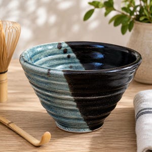 May include: A ceramic matcha bowl featuring a ribbed exterior with a split color design. One half of the bowl is finished in a glossy, speckled light blue glaze, while the other half is finished in a deep, solid black glaze with a smooth texture.