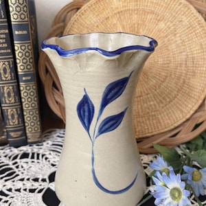 May include: A beige ceramic vase with a ruffled rim and blue accents. The vase features a blue leaf design and is approximately 8 inches tall. Decorative vase for flowers or home decor.