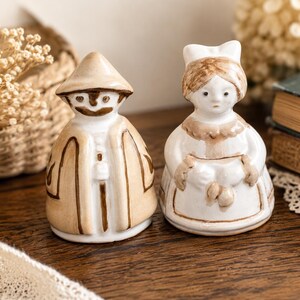 May include: Two ceramic figurines, a man and a woman, stand side-by-side on a wooden surface. The man wears a tan coat and hat, while the woman is dressed in a white dress and apron. The figurines are hand-painted with brown accents.