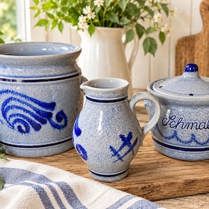 May include: A set of three light blue salt-glazed stoneware pieces featuring dark blue hand-painted patterns. The collection includes a large crock, a small pitcher marked 0.2L, and a lidded jar with the word Schmalz painted on the front.