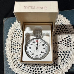 May include: A vintage stopwatch in its original box, labeled "SWISS MADE." The stopwatch has a white face with black numerals and a red hand. The box is open, revealing the watch nestled inside. The watch is on a white lace doily.