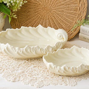 May include: Two cream-colored ceramic bowls shaped like large leaves with ruffled edges and a curled handle on one side. The bowls have a glossy finish and are arranged on a white lace doily, showcasing a textured, organic design.