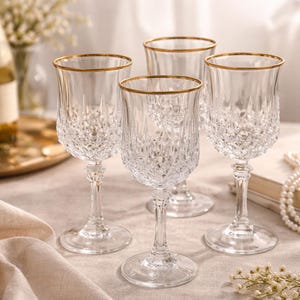 Op de afbeelding: Vier heldere kristallen wijnglazen met gouden randen. De glazen hebben een diamantgeslepen patroon en een delicate steel. Ze staan op een lichtgekleurd oppervlak.