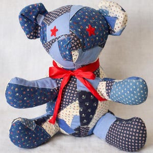 Könnte beinhalten: Ein handgefertigter Patchwork-Teddybär in Blau- und Weißtönen mit einem roten Band um den Hals. Der Bär hat eine Vielzahl von floralen und gemusterten Stoffen mit roten Sternen auf dem Kopf. Ein charmanter, handgefertigter Plüschtier.
