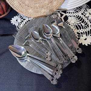 Puede incluir: Una colección de cubiertos de acero inoxidable que incluye varias cucharas y tenedores dispuestos sobre una superficie circular de piedra gris. Cada pieza presenta un diseño de mango decorativo y ornamentado con un motivo texturizado en abanico.