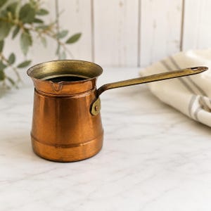 May include: A copper coffee pot with a wide, flared rim and a small pouring spout. The pot features a long, flat brass handle attached to the side with a single rivet. The body of the pot has a smooth, metallic finish with visible signs of age.