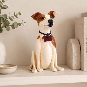 May include: A sitting dog figurine with white and brown fur, featuring a black collar and a dark red bone-shaped tag. The dog has pointed ears, a black nose, and a smooth finish, depicted in a seated position with its front paws placed forward.