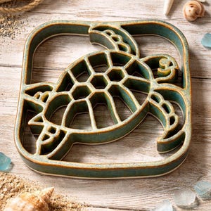 May include: A teal ceramic turtle-shaped trivet with a square frame. The trivet has a detailed turtle design with a hexagonal pattern on its shell. The background includes a starfish, seashells, and sand, suggesting a coastal theme.
