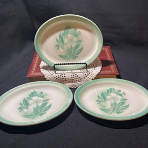 May include: A set of three oval serving dishes. The dishes are white with a light green rim and a floral design in the center. The largest dish is on a black metal stand. The dishes are arranged on a dark surface.