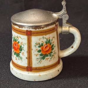 May include: A small, ceramic beer stein with a hinged metal lid. The stein is decorated with orange roses and green leaves on a white background, framed by brown and red accents. It has a white handle.