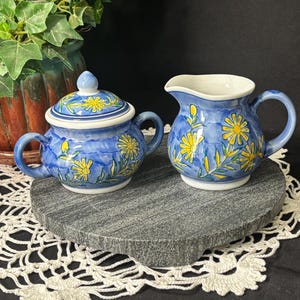 May include: A blue ceramic sugar bowl with a lid and a matching creamer. Both pieces are decorated with yellow daisy-like flowers and green leaves. The sugar bowl has a small handle on each side, and the creamer has a single handle.