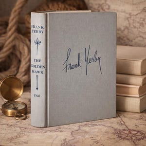 May include: A hardcover book titled "The Golden Hawk" by Frank Yerby. The book has a light gray cover with blue text and a signature. A compass and rope are in the background, suggesting a nautical theme.