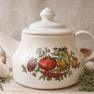 May include: Cream-colored ceramic teapot with a lid and handle. The teapot is decorated with a colorful illustration of vegetables, including a red tomato, green pepper, and orange carrot. Garlic and herbs are arranged nearby.