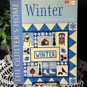 Puede incluir: Un libro titulado "Winter" de Lois Krushina Fletcher. La portada presenta un diseño de edredón azul y blanco con ilustraciones temáticas de invierno, incluyendo un muñeco de nieve, una casa y copos de nieve. Es parte de la serie "The Quilter's Home".