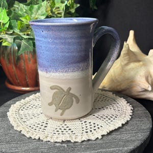 May include: A ceramic mug with a blue and white glaze. The mug has a turtle design and a curved handle. The mug is on a white lace doily. The mug is approximately 10 cm tall.