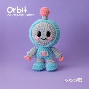 Puede incluir: Un juguete robot amigurumi de crochet llamado Orbit. El robot es azul y gris con detalles en rosa. El robot tiene una cara amigable con ojos negros y una sonrisa.