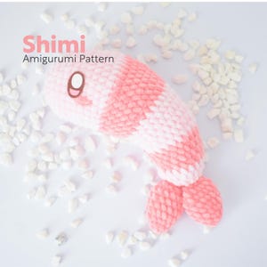 May include: A handmade amigurumi shrimp toy in pink and white, with a brown eye and a tail. The text "Shimi Amigurumi Pattern" is in the upper left corner. The toy is surrounded by small white stones.
