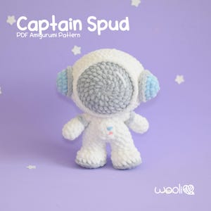 以下が含まれることがあります： 白い毛糸で編まれた、宇宙飛行士のアミグルミ人形。顔はグレーで、耳はブルーのディテール。人形の名前は「キャプテン・スパッド」で、PDFのアミグルミパターンです。