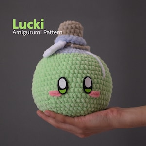 May include: A green crocheted amigurumi toy shaped like a bottle with a brown and blue crocheted stopper. The toy has a cute face with black eyes and pink cheeks. The toy is called "Lucki" and is an amigurumi pattern.