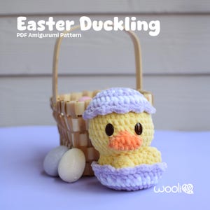 Puede incluir: Un patrón de amigurumi de patito de Pascua tejido a crochet. El patito lleva una cáscara de huevo tejida a crochet de color morado y blanco. El patito está sentado frente a una cesta de mimbre con huevos de Pascua.