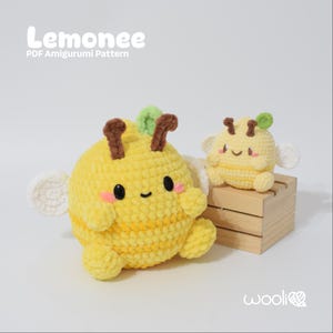 May include: Two yellow crocheted amigurumi bees with smiling faces, brown antennae, and white wings. The larger bee has pink cheeks and is next to a smaller bee on a wooden box. The text "Lemonnee PDF Amigurumi Pattern" is at the top.