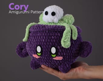 Può includere: Un calderone di uncinetto viola con un coperchio verde e un fantasma di uncinetto bianco in cima. Il calderone ha un viso carino con occhi verdi e guance rosa. "Cory Amigurumi Pattern" è scritto in bianco su uno sfondo viola.