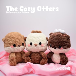 May include: Three crocheted otter plushies in shades of brown and cream sit on a pink cloth. Each otter has a round head, small ears, and a white face with black eyes and pink cheeks. The text "The Cozy Otters" is at the top.