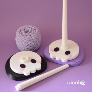May include: A craft-themed image featuring a purple ball of yarn and two yarn holders. The yarn holders have a skull design with black eyes and pink cheeks. One holder is on a black base, the other on a purple base. A white rod and a white tube are also present.