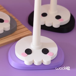 May include: Close-up of three decorative skull-shaped stands. The skulls are white with black eyes and pink cheeks, set on purple, grey, and black bases. The stands have white poles. The background is a solid purple.
