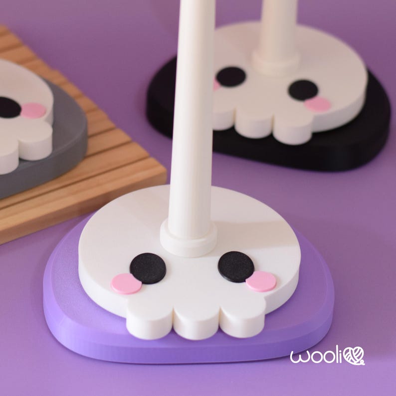 May include: Close-up of three decorative skull-shaped stands. The skulls are white with black eyes and pink cheeks, set on purple, grey, and black bases. The stands have white poles. The background is a solid purple.