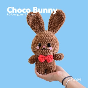 Może przedstawiać: Brązowy królik szydełkowy z czerwoną muszką. Królik jest trzymany w dłoni. Tekst "Choco Bunny PDF Amigurumi Pattern" znajduje się w górnej części obrazu.