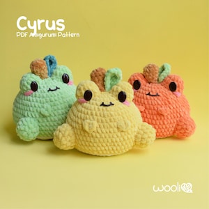 May include: Three crocheted frog plushies in green, yellow, and orange. The plushies have black eyes and pink mouths. The plushies are sitting on a yellow surface. The text "Cyrus PDF Amigurumi Pattern" is at the top of the image.