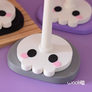 May include: Close-up of three decorative skull-shaped stands. Each stand features a white skull design with black eyes and pink cheeks. The stands are on grey, black, and purple bases, with a white pole extending upwards.