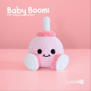 Könnte beinhalten: Ein rosa und weiß gehäkeltes Amigurumi-Spielzeug in Form einer Babyflasche. Das Spielzeug hat ein lächelndes Gesicht mit schwarzen Augen und ein rosa-weiß gestreiftes Oberteil. Das Spielzeug sitzt auf einer rosa Oberfläche. Der Text "Baby Boomi PDF Amigurumi Pattern" befindet sich oben im Bild.