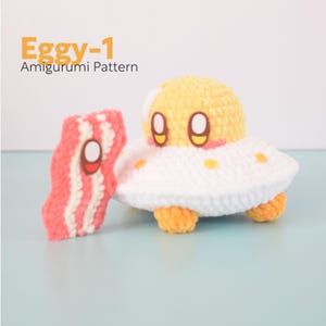 May include: A crocheted amigurumi pattern featuring a yellow and white egg-shaped character with a smiling face, resembling a UFO. Beside it is a crocheted piece of bacon. The text "Eggy-1 Amigurumi Pattern" is in the upper left corner.