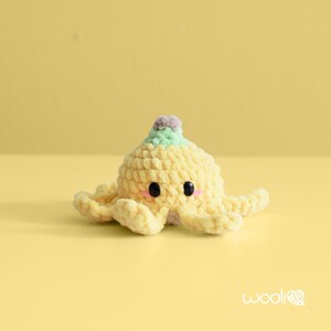 May include: A handmade, yellow crocheted octopus with black eyes and pink cheeks. The octopus has a light green and gray top and eight tentacles. The background is a solid yellow.