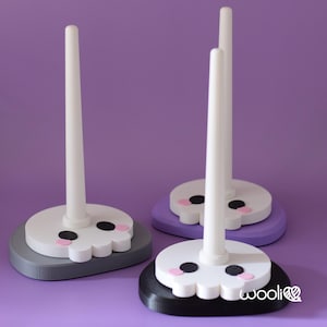 May include: Three decorative yarn holders with a skull design. Each holder has a white, cylindrical post attached to a base. The bases are in grey, purple, and black, with a white skull face, black eyes, and pink cheeks. The word "wooli" is printed on the black base.