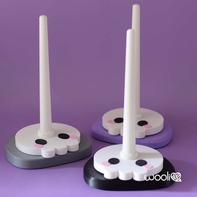 May include: Three decorative yarn holders with a skull design. Each holder has a white, cylindrical post attached to a base. The bases are in grey, purple, and black, with a white skull face, black eyes, and pink cheeks. The word "wooli" is printed on the black base.