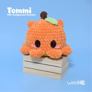 Puede incluir: Un amigurumi de pulpo de ganchillo naranja con ojos negros, mejillas rosas y un tallo marrón con una hoja verde. El pulpo está sentado en una pequeña caja de madera. El texto "Tommi PDF Amigurumi Pattern" está en la parte superior.
