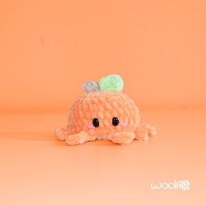 May include: A plush orange octopus with black eyes and pink cheeks. The octopus has a small green and gray accent on top. The background is a solid orange color. The word "wooli" is in the bottom right corner.