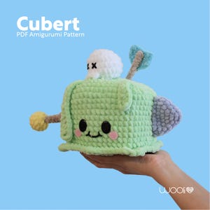 Può includere: Un giocattolo amigurumi di uncinetto verde a forma di cubo con una nuvola bianca di uncinetto in cima. Il giocattolo ha un viso amichevole con occhi neri e guance rosa. Il giocattolo si chiama "Cubert" ed è uno schema amigurumi PDF.
