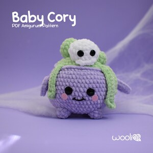 May include: A crocheted amigurumi toy named "Baby Cory" wearing a green and white ghost hat. The toy is purple and has a friendly face with black eyes and a pink smile. The text "PDF Amigurumi Pattern" is also visible.