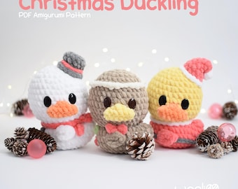 May include: Three crocheted ducklings with black eyes and orange beaks. The left is white with a gray hat and pink scarf, the center is tan with a pink bow tie, and the right is yellow with a pink hat. Text reads: Christmas Duckling PDF Amigurumi Pattern.