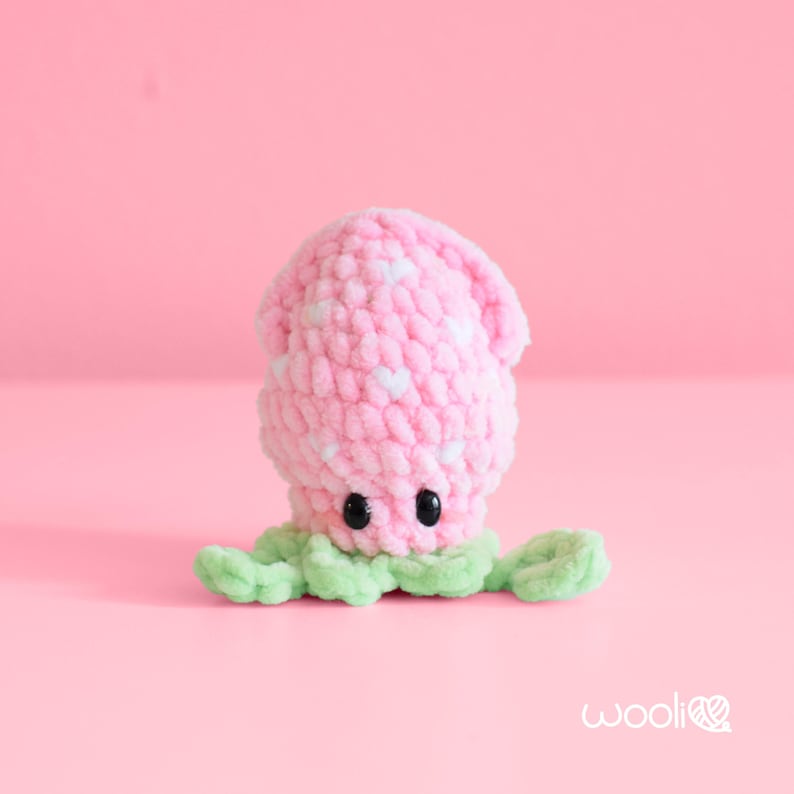 May include: A handmade crochet octopus toy in pink with white polka dots. The octopus has black button eyes and green tentacles. The background is a solid pink.