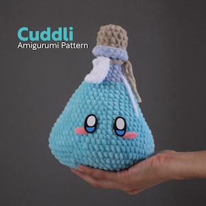 May include: A blue crocheted amigurumi bottle with a brown cork top and a white crocheted label. The bottle has a cute face with black eyes and pink cheeks.