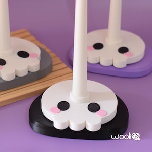 May include: Close-up of three white skull-shaped yarn holders with black eyes and pink cheeks. Each holder has a white pole and sits on a different coloured base: black, grey, and purple. The bases are round and the background is purple.