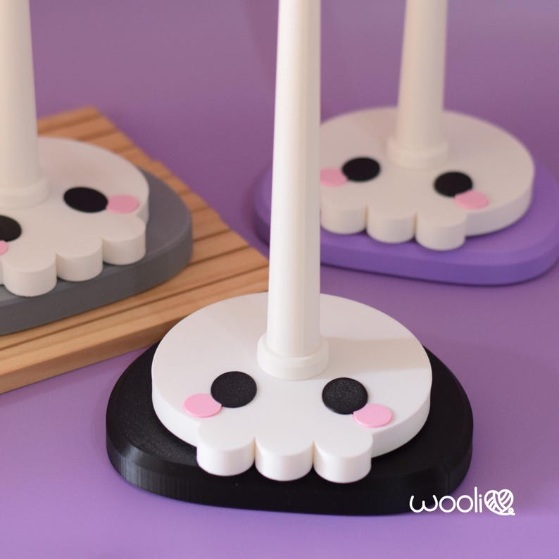 May include: Close-up of three white skull-shaped yarn holders with black eyes and pink cheeks. Each holder has a white pole and sits on a different coloured base: black, grey, and purple. The bases are round and the background is purple.