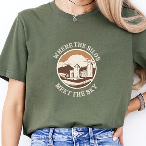 Peut inclure: Un t-shirt vert avec un graphique circulaire brun représentant un coucher de soleil, trois silos et le texte "Where the Silos Meet the Sky".
