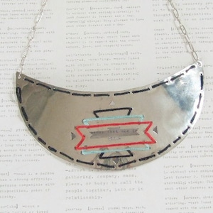 Puede incluir: Collar de metal plateado con un colgante en forma de media luna. El colgante presenta costuras negras y diseños geométricos bordados en rojo, turquesa y negro. El texto "that one is" es visible. Una joya única.