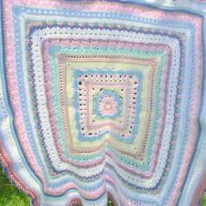 May include: A handmade, square crochet blanket in pastel shades. The blanket features concentric squares of pink, blue, yellow, and white, with intricate stitch patterns and a scalloped edge. The blanket is made of soft yarn.
